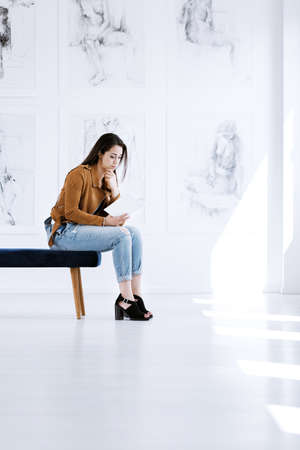 Young woman sitting on stool and reading guidebook while visiting art gallery