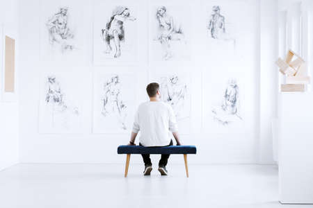 Man relaxing in art gallery while sitting on stool in front of white wall with drawings. Art gallery conceptの写真素材