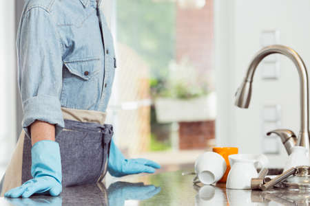 Cropped image of woman in apron wearing cleaning rubber gloves standing at the kitchen sink full of...