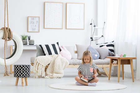 Girl in striped shirt reads book on white round carpet in kid room with tire on lineの写真素材