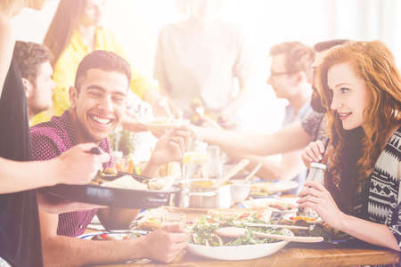 Happy couple eating healthy lunch at vegan restaurant with group of friends, beautiful girl drinking water, smiling boy chatting with waitressの写真素材
