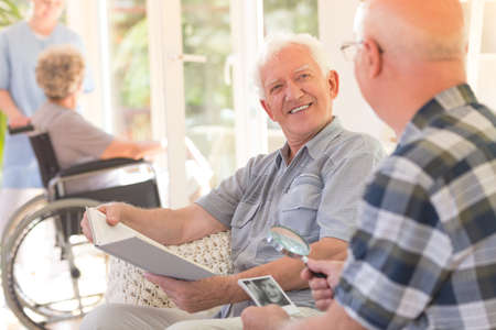 Smiling grandpa sitting with a book and talking to his elder friend at the rest houseの写真素材