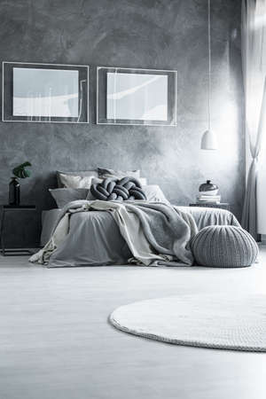 Simple posters on dark wall above king-size bed with knot pillow in bedroom with pouf and white carpetの写真素材