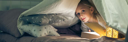 Mysterious girl hiding under the covers with book and torchの写真素材