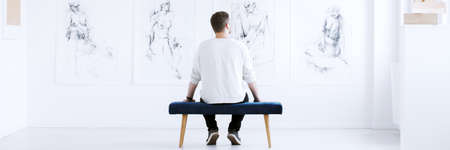 Young man sitting back on the bench alone looking at collection of charcoal drawings hanging on white wall in bright art centerの写真素材