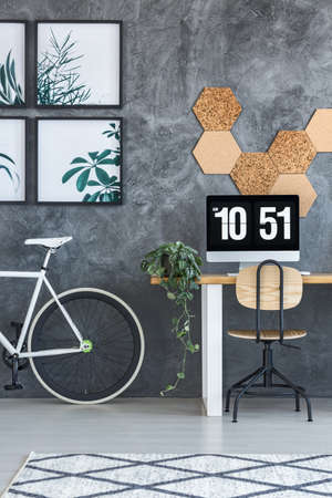 Monitor and plant on desk with classic chair in dark work area with bicycle and posters on textured wallの写真素材