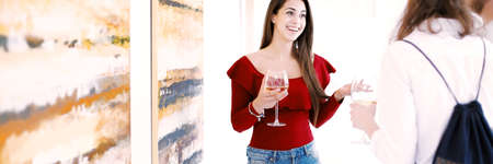 Happy female artist drinking wine and talking with audience at her own exhibition of paintings in art galleryの写真素材