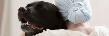 Back view of sick person with blue headscarf hugging black dog during animal assisted therapy