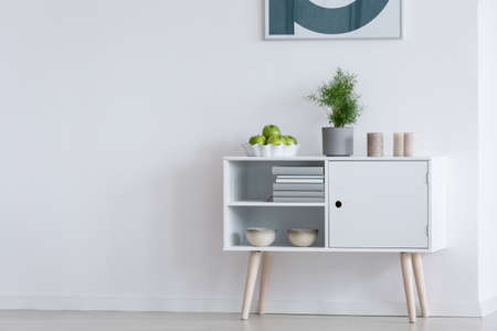 Apples in bowl, plant in grey pot and candles on white modern cupboard against white wallの写真素材