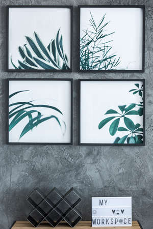 Simple square paintings with leaves hanging on raw wall above table with wine rack and handmade posterの写真素材