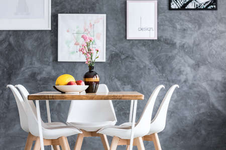 Fruits in white bowl and flowers in decorative vase on dining table with white chairs against...