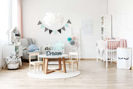 Fabric pennant banner hanging over a child's white cradle standing next to a large armchair with a blue, cloud shaped pillow looking in the direction of wooden chest of drawersの写真素材