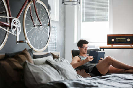 Man sitting on chaise lounge and reading book next to retro radio in bedroom with bike on wallの写真素材