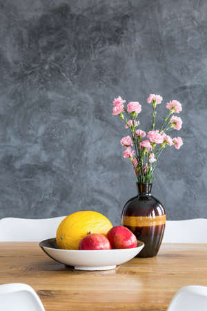 Pomegranate and melon in white bowl and pink flowers in decorative vase on wooden dining tableの写真素材