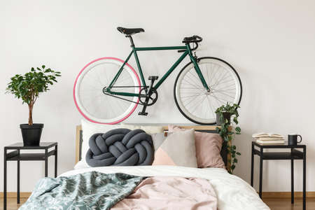 Black bike on bedhead in bedroom with pink and green blankets on bed and plant on cabinetの写真素材