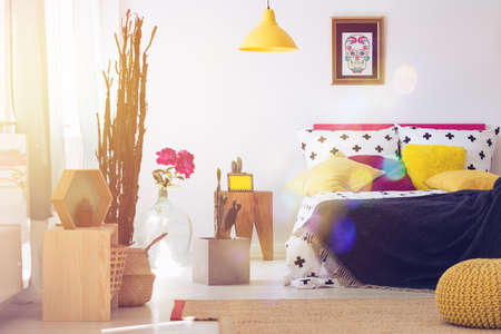 Yellow pouf and lamp in mexican bedroom with calavera skull poster, pink flowers, wooden stool and other furnitureの写真素材