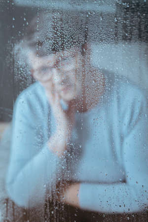 Photo through the window with raindrops with sad grandmother in background