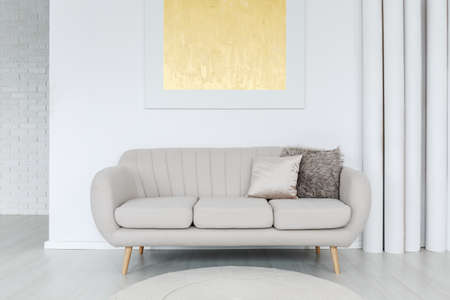 Gold painting on the wall above grey sofa with pillows in bright living room interior with tube decorationの写真素材