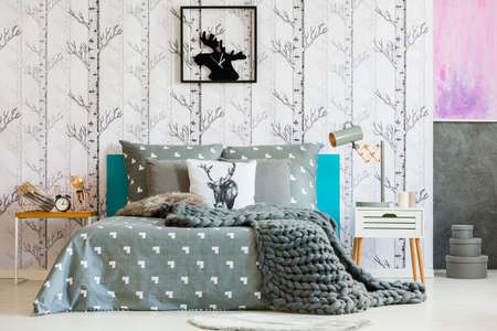 Grey blanket on king-size bed and lamp on nightstand in bright bedroom with forest motif