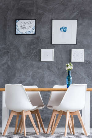 White chairs at dining table with flowers in blue vase in room with paintings on dark concrete wall