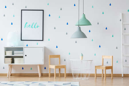 Poster above white cupboard in bright kid's room with white designer table and wooden chairsの写真素材