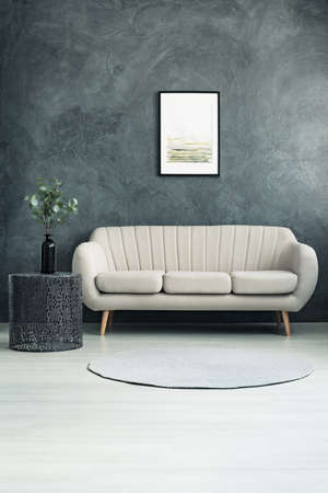 Bright couch standing in living room with grey textured wall and twigs with leaves in a glass vase on metal tableの写真素材
