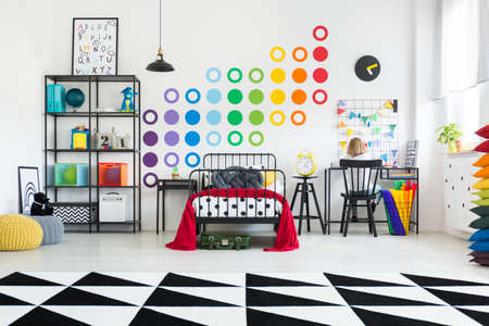 Geometric carpet in spacious bright kid's room with red bedding on the bed, toys on a shelf and black chair at a deskの写真素材