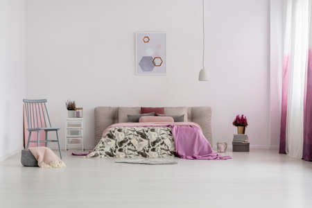 Patterned bedding on king-size bed and grey chair in bright bedroom with poster on the wall