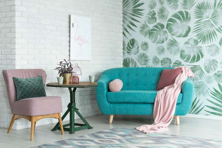 Green table with a plant between pink chair and blue sofa in floral living room with wallpaper and poster