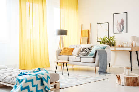 Patterned blanket on settee and candle on wooden stump in spacious living room with yellow curtains and beige sofaの写真素材