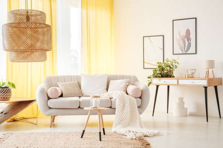 Rattan lamp and wooden stool on carpet in warm bohemian living room with yellow curtains and beige settee with white pillowsの写真素材