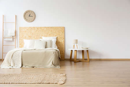 Lamp on wooden table next to bed with pillows in natural bedroom with ladder, paper clock and rugの写真素材