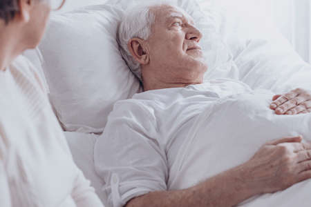 Woman visiting senior man sick with cancer lying in bed at the hospitalの写真素材