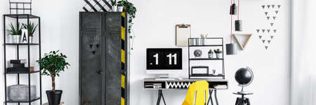 Black globe standing next to a wooden desk and yellow chair in bright interior for a freelancerの写真素材