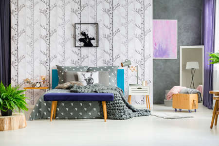Moose clock above king-size bed with grey bedsheets in forest bedroom with fern, stool and wooden boxの写真素材