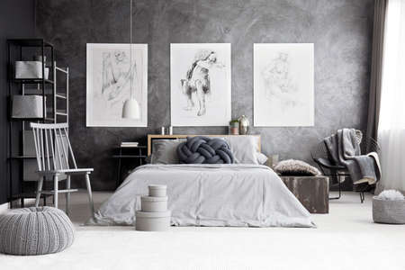 Pouf and boxes near grey chair in monochromatic bedroom with drawings on concrete wall above king-size bedの写真素材