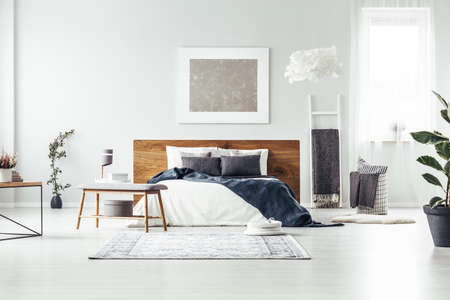 Carpet and wooden bench near king size bed with navy blue blanket in spacious bedroom with silver paintingの写真素材