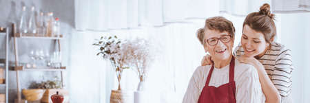 Panoramic view of happy loving grandmother hugged by adult supporting granddaughter while baking together in cozy kitchenの写真素材