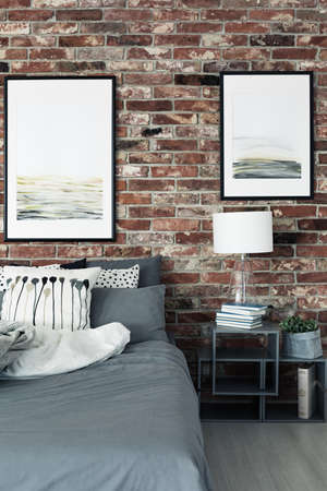 Paintings on brick wall above bed and cabinet with lamp in modern bedroom interiorの写真素材