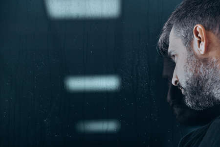 Caucasian man with gray hair watching rain through window at nightの写真素材