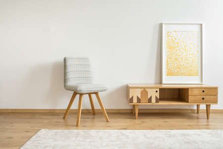 Grey chair next to wooden cupboard with gold painting in room with white wall with copy spaceの写真素材