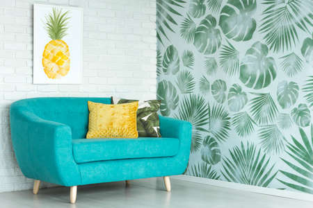 Yellow pillow on blue sofa against a wall with pineapple poster in living room with floral wallpaperの写真素材