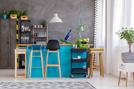 Plant on white cabinet near blue kitchenette, white lamp, wooden stools and black cabinet in room with textured wall