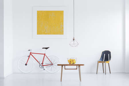 Lamp above wooden table with fruits in white apartment with gold painting above red bike and designer chairの写真素材