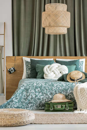 Rattan lamp above bed with hat and white pillow in traveler's bedroom with suitcase and pouf