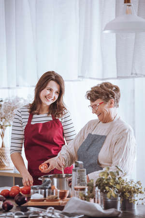 Granddaughter holding tomato and grandmother mixing italian soup in the kitchenの写真素材