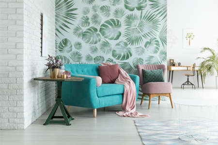 Green table with plant next to blue sofa and pink armchair with pillow against green wallpaper in living room