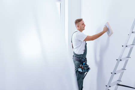 Young smiling painter with tool belt and workwear smoothing a wall before painting