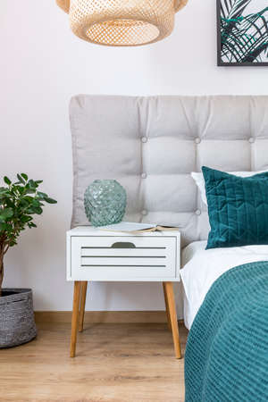 Close-up of glass vase and book on white nightstand between bed with green pillow and plant in simple bedroom interiorの写真素材