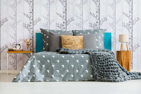 Two bedside tables with lamps, clock and book standing by the bed with grey bedclothes and golden cushionの写真素材